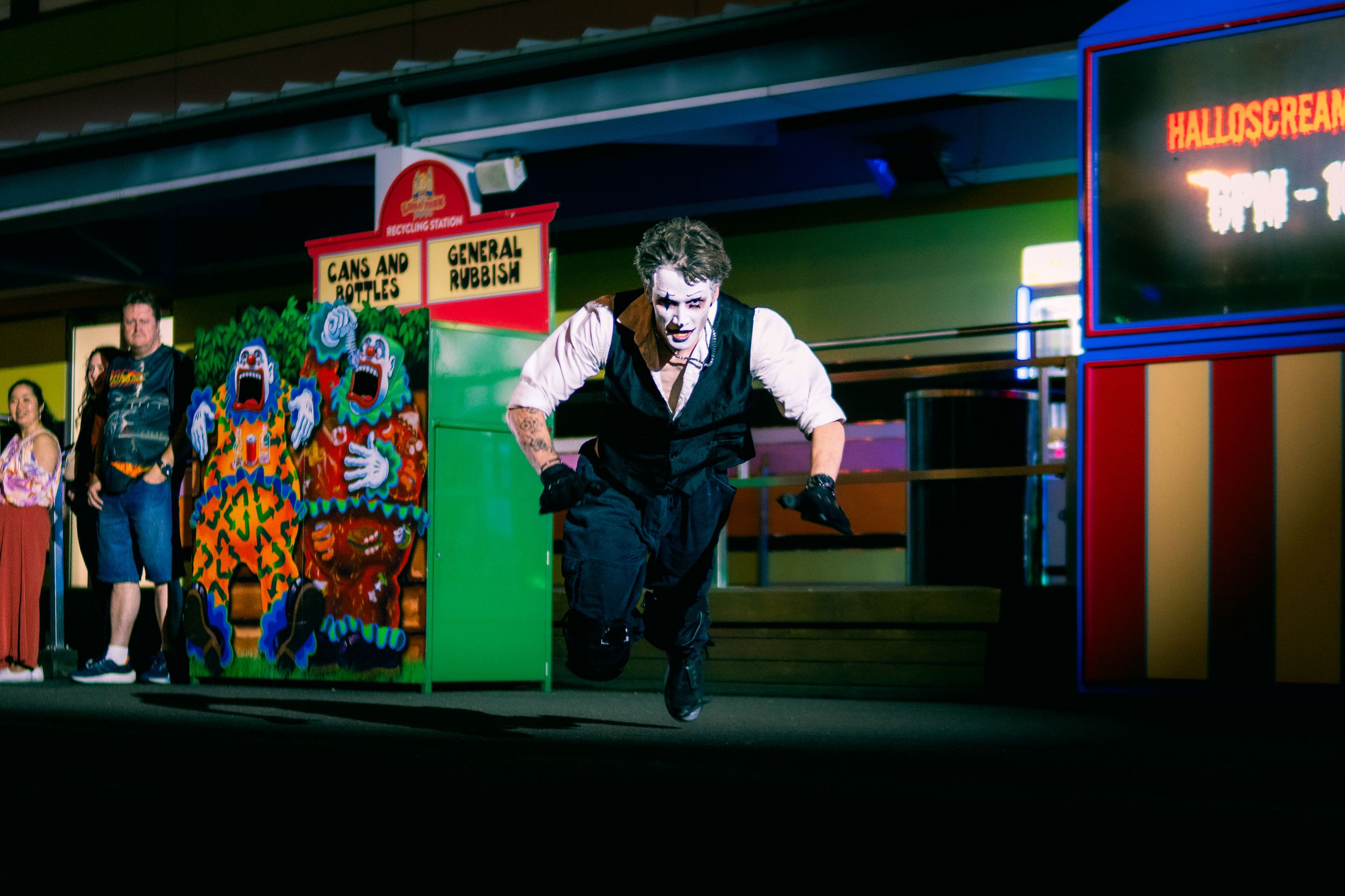 Man in zombie makeup runs past colorful carnival game booths at Halloscreen Halloween event.