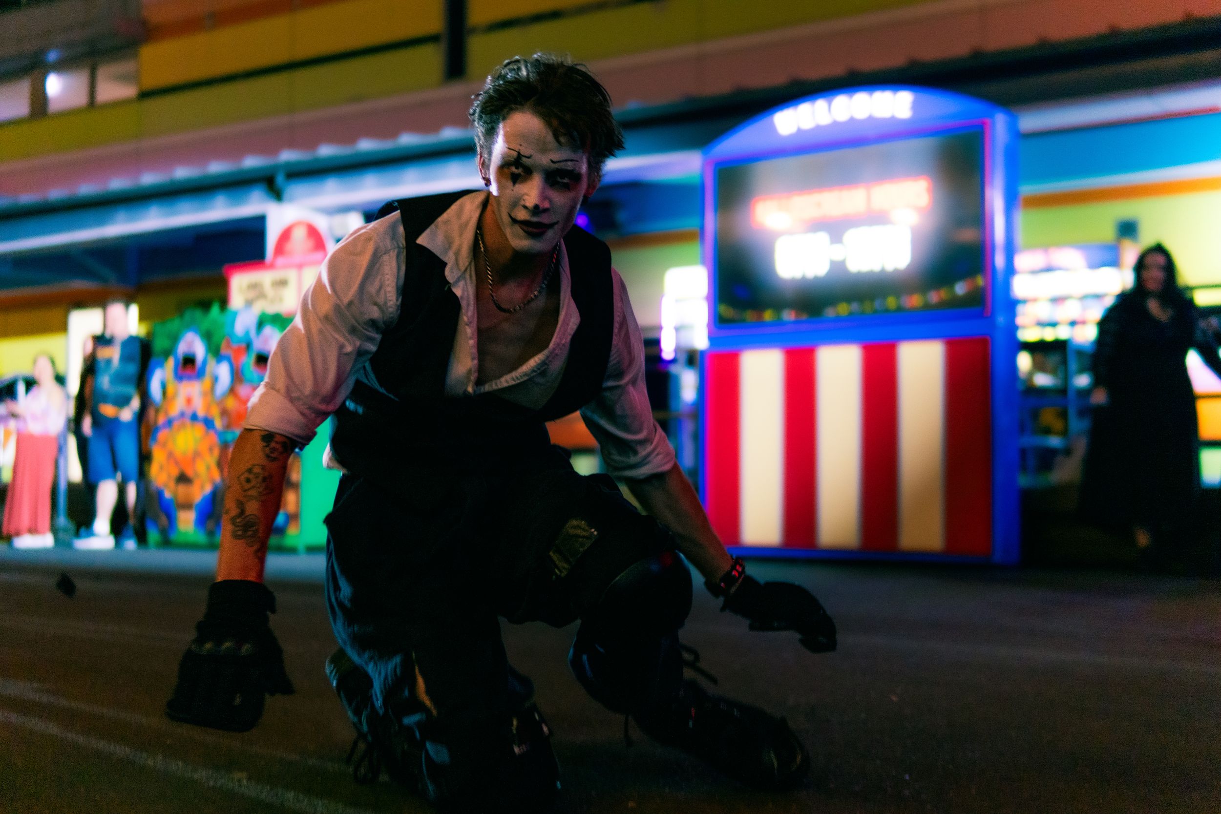 Male zombie performer in vest and face paint crouches aggressively at colorful Halloween carnival.
