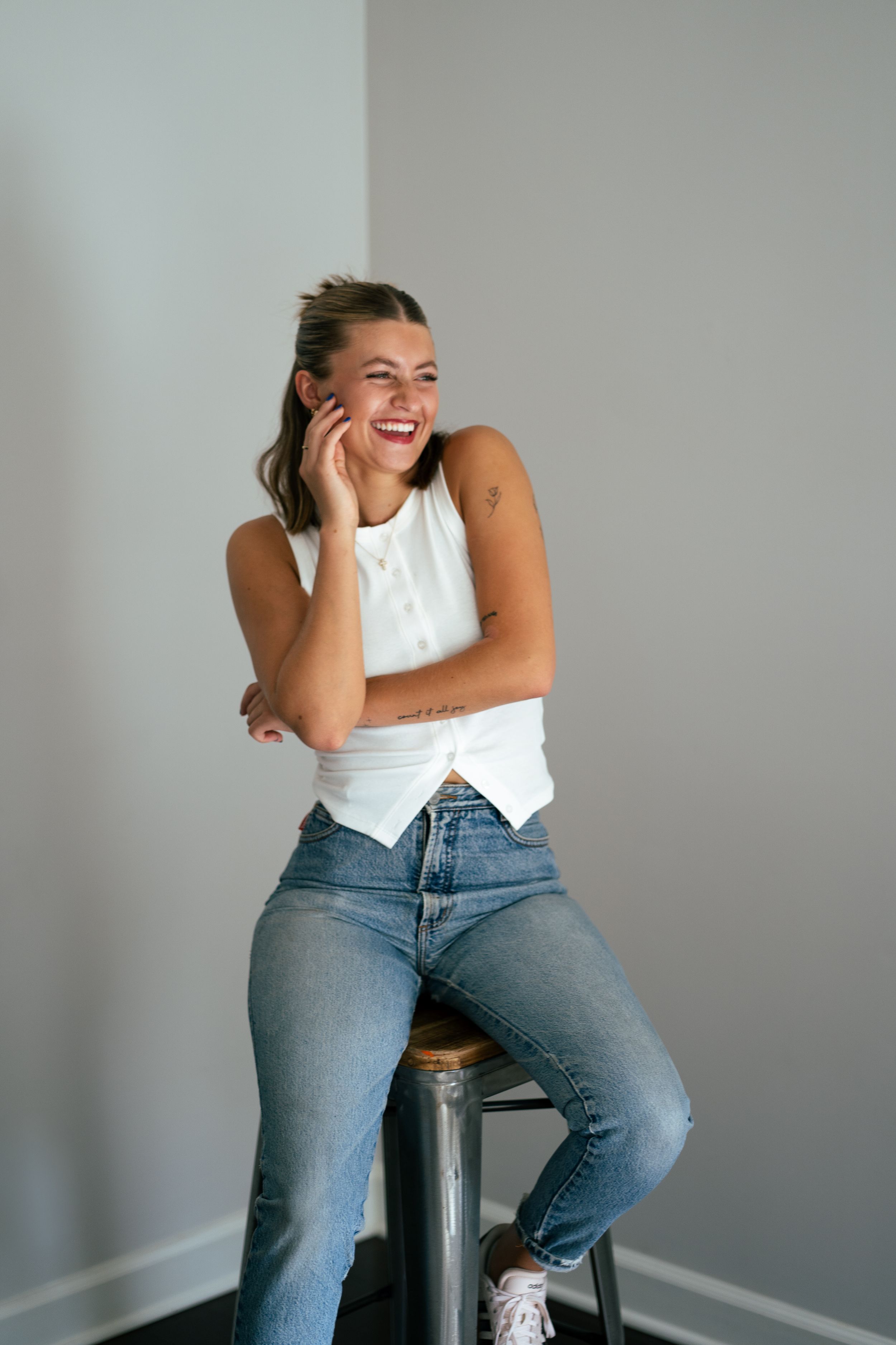Smiling young woman in white top and jeans sitting on a stool, laughing with hand near face.
