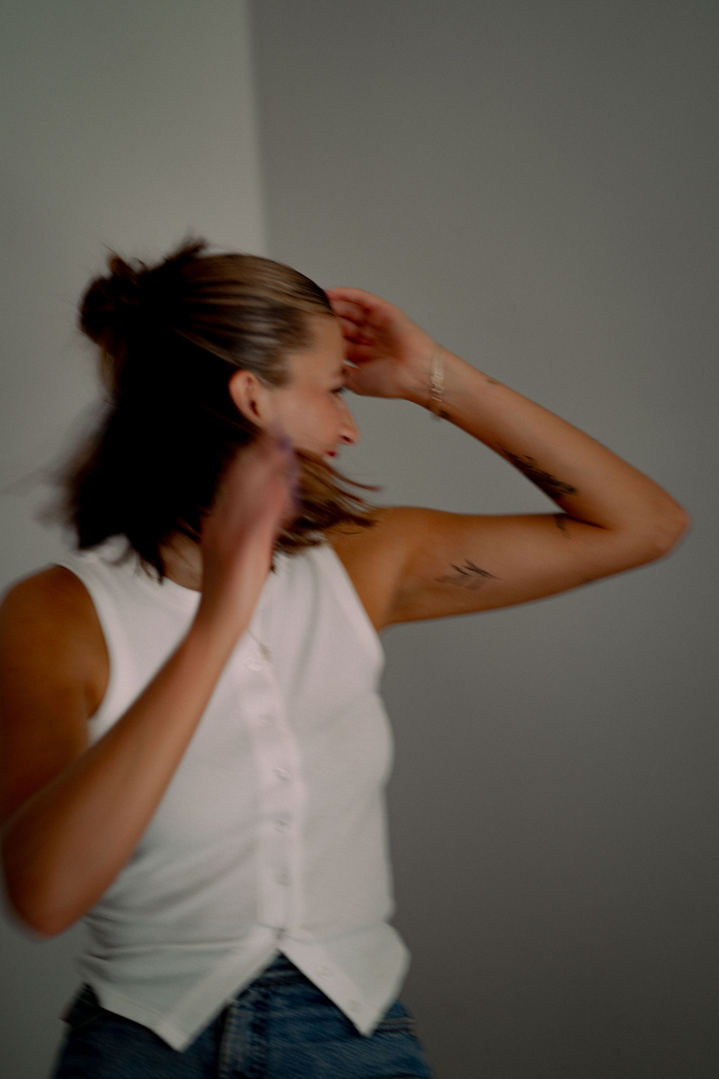 Woman in white sleeveless vest and jeans dancing with motion blur, tattoo visible on upper arm.