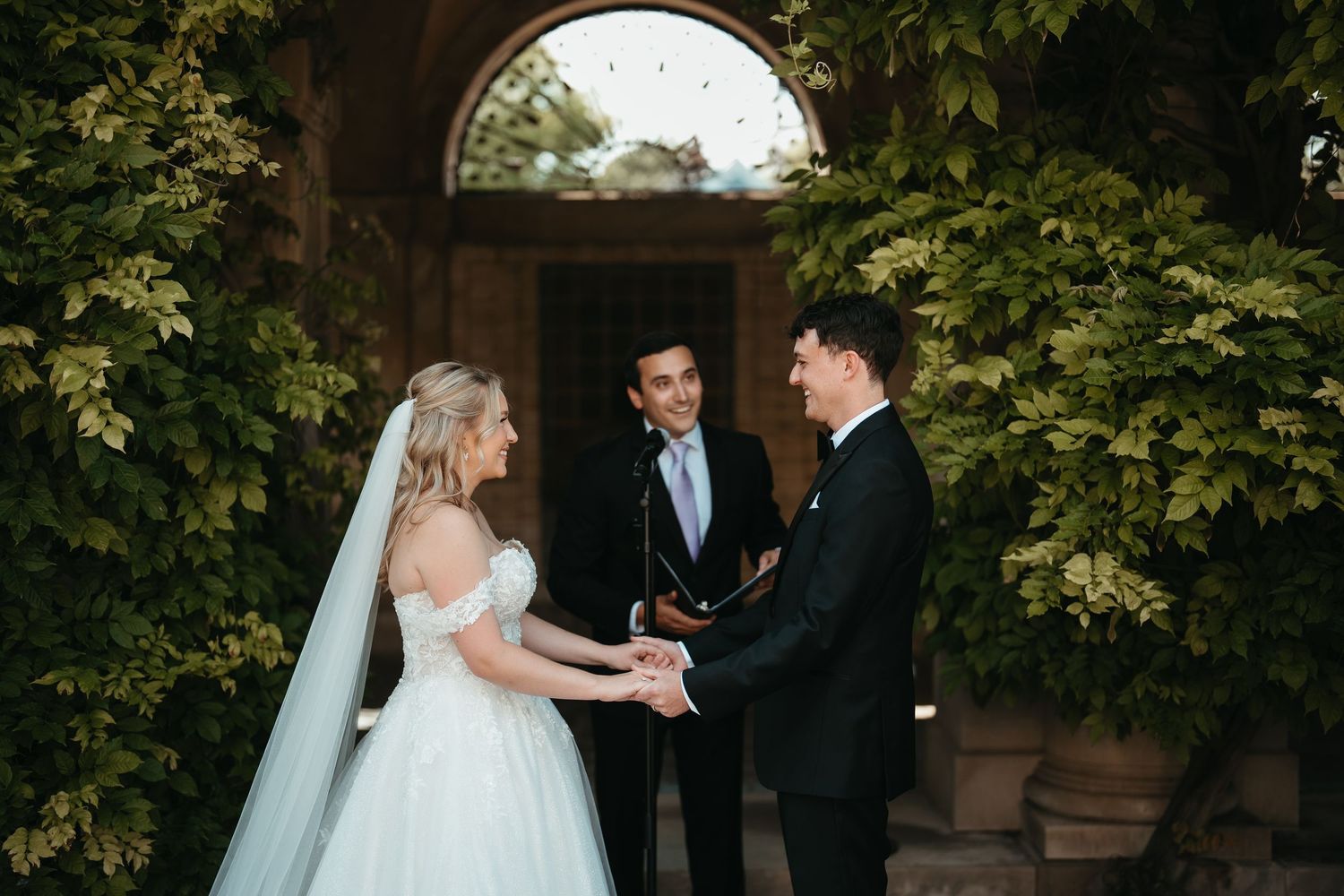 Bride and groom hold hands during outdoor wedding ceremony with officiant, surrounded by lush green ivy walls.