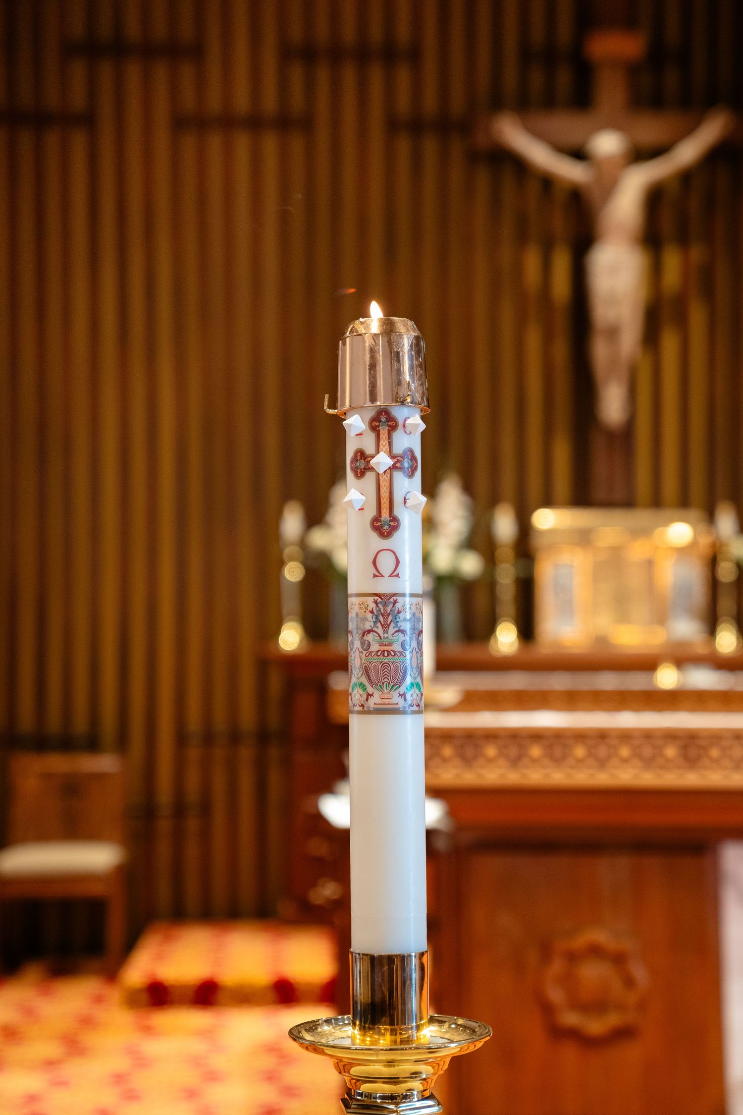 Lit paschal candle with religious symbols on golden stand in Catholic church with crucifix altar in background.