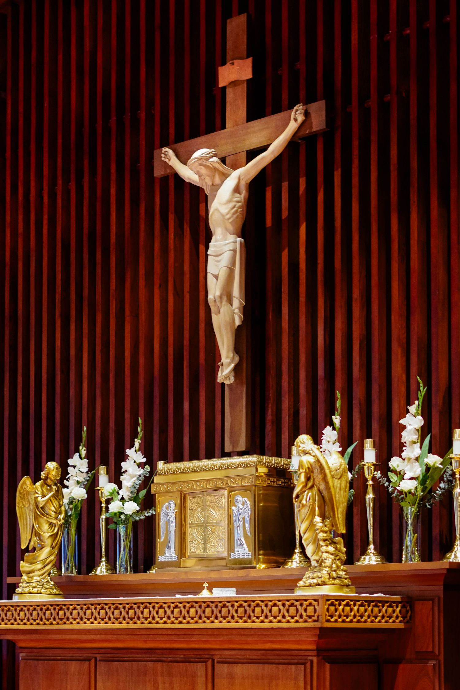 Catholic church altar with crucifix, golden angel statues, and white floral arrangements against rich wood paneling.