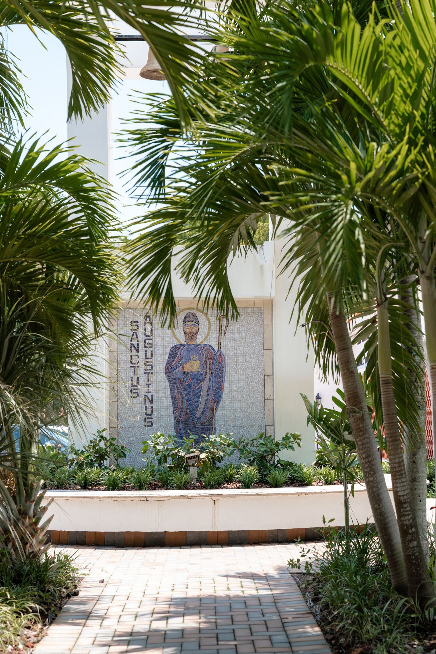 Mosaic mural of Sanctus Augustinus on a white wall surrounded by lush tropical palm trees and a brick pathway.