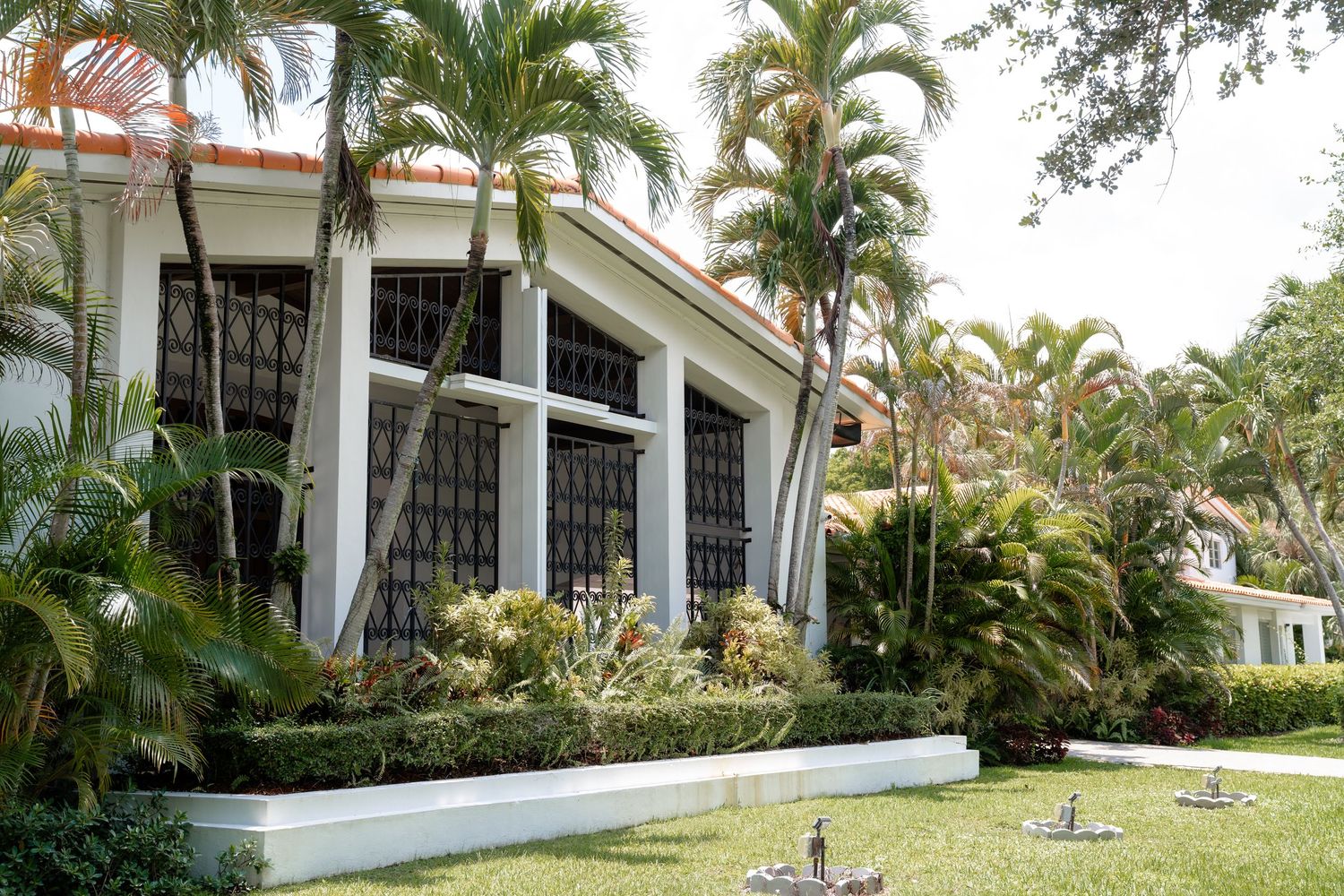 White tropical villa with ornate black window grilles surrounded by lush palm trees and manicured hedges in a sunny garden.