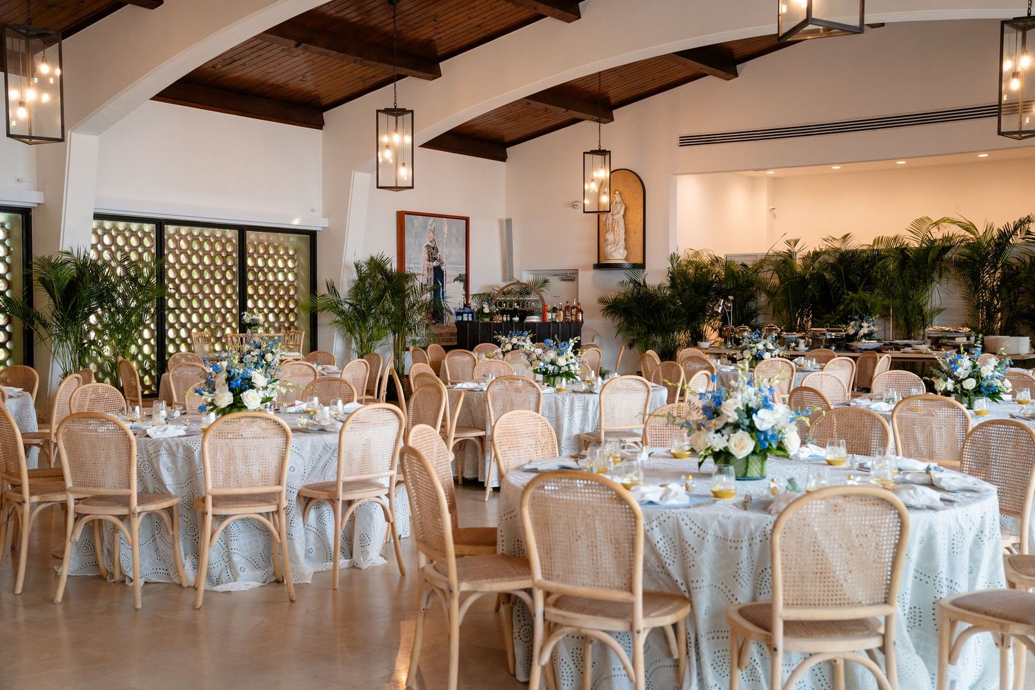 Elegant wedding reception hall with round tables, wicker chairs, floral centerpieces, and tropical greenery under wood beam ceilings.