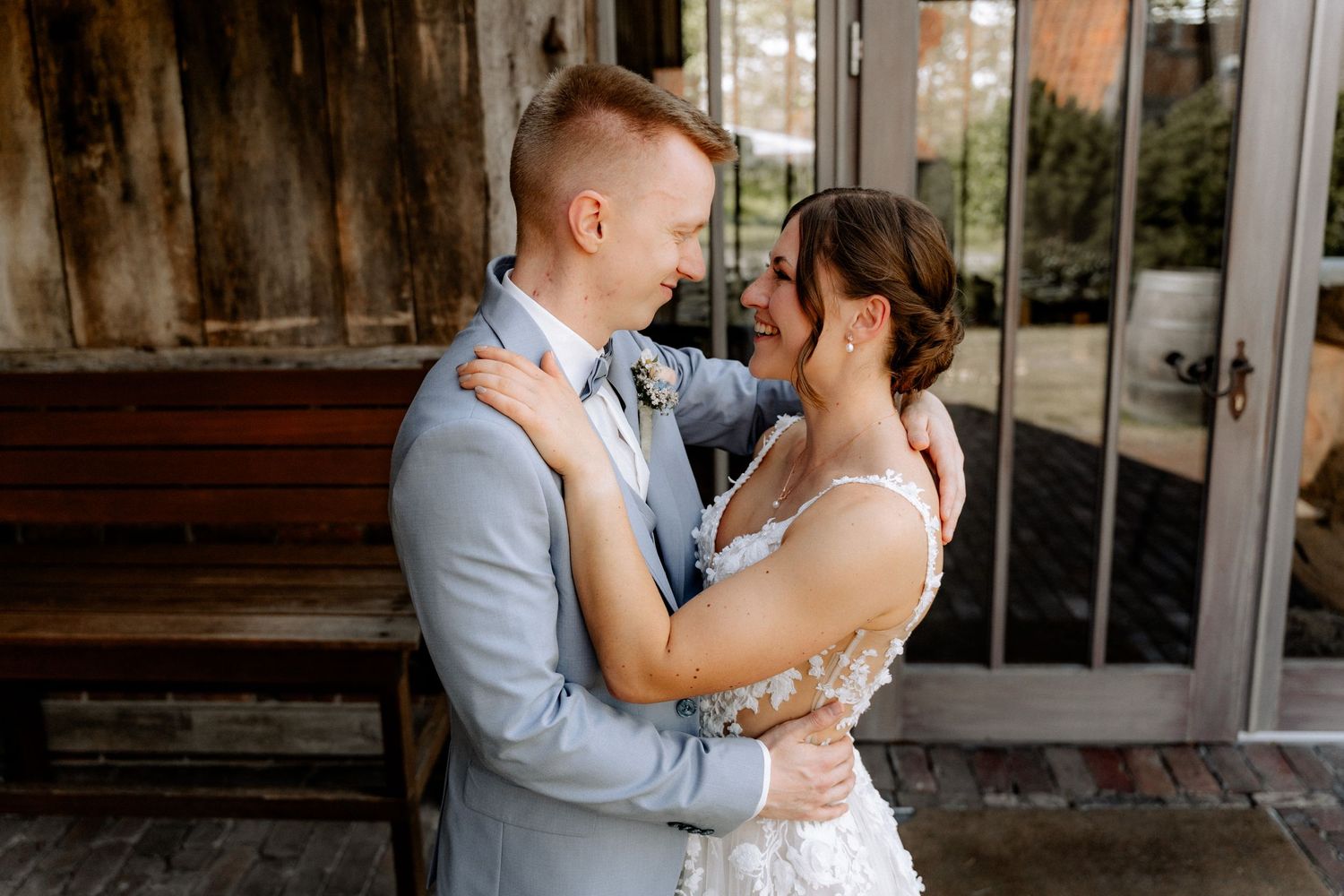 A joyful bride and groom share a romantic embrace, smiling at each other outside a rustic wooden venue.