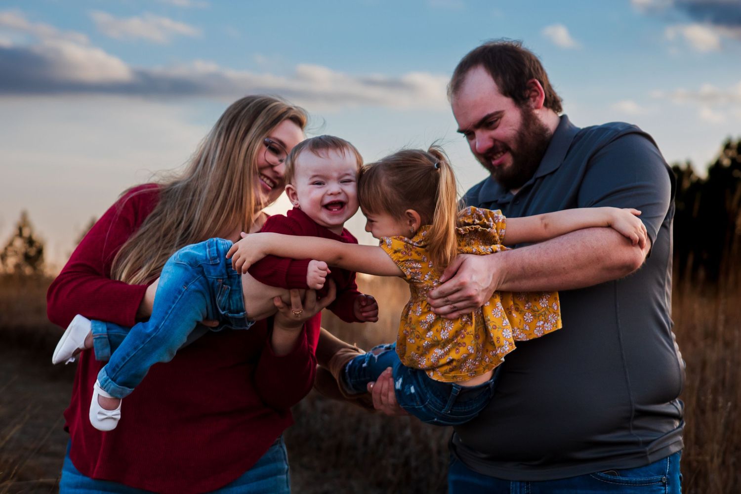 Happy family of four playing outdoors at sunset, parents swinging laughing toddlers in a field.