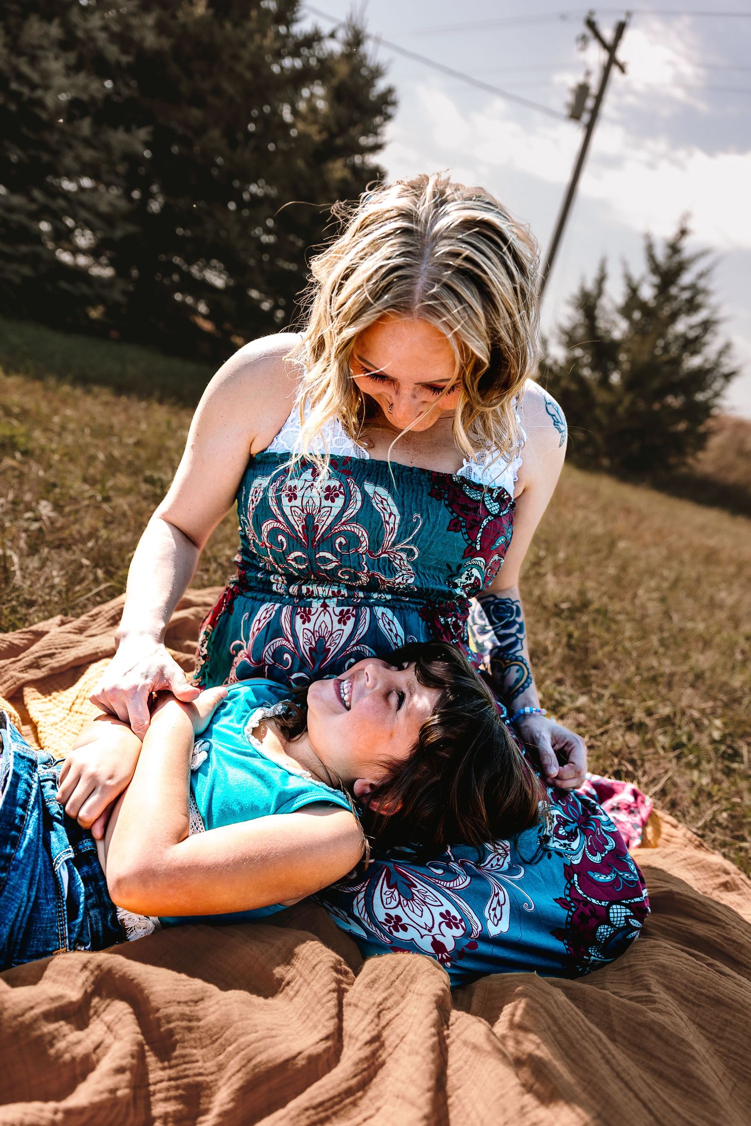 A woman in a floral dress sits outdoors on a blanket with a child lying in her lap, sharing a joyful moment.
