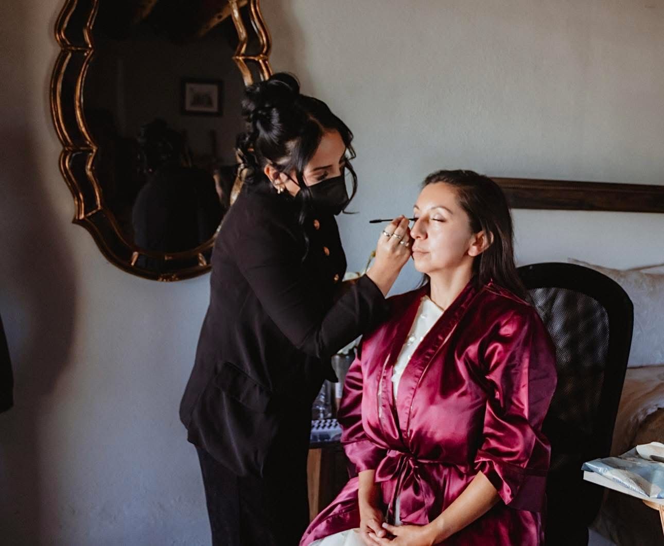 Makeup artist in black mask applying eye makeup to a bride in a burgundy satin robe before her wedding.