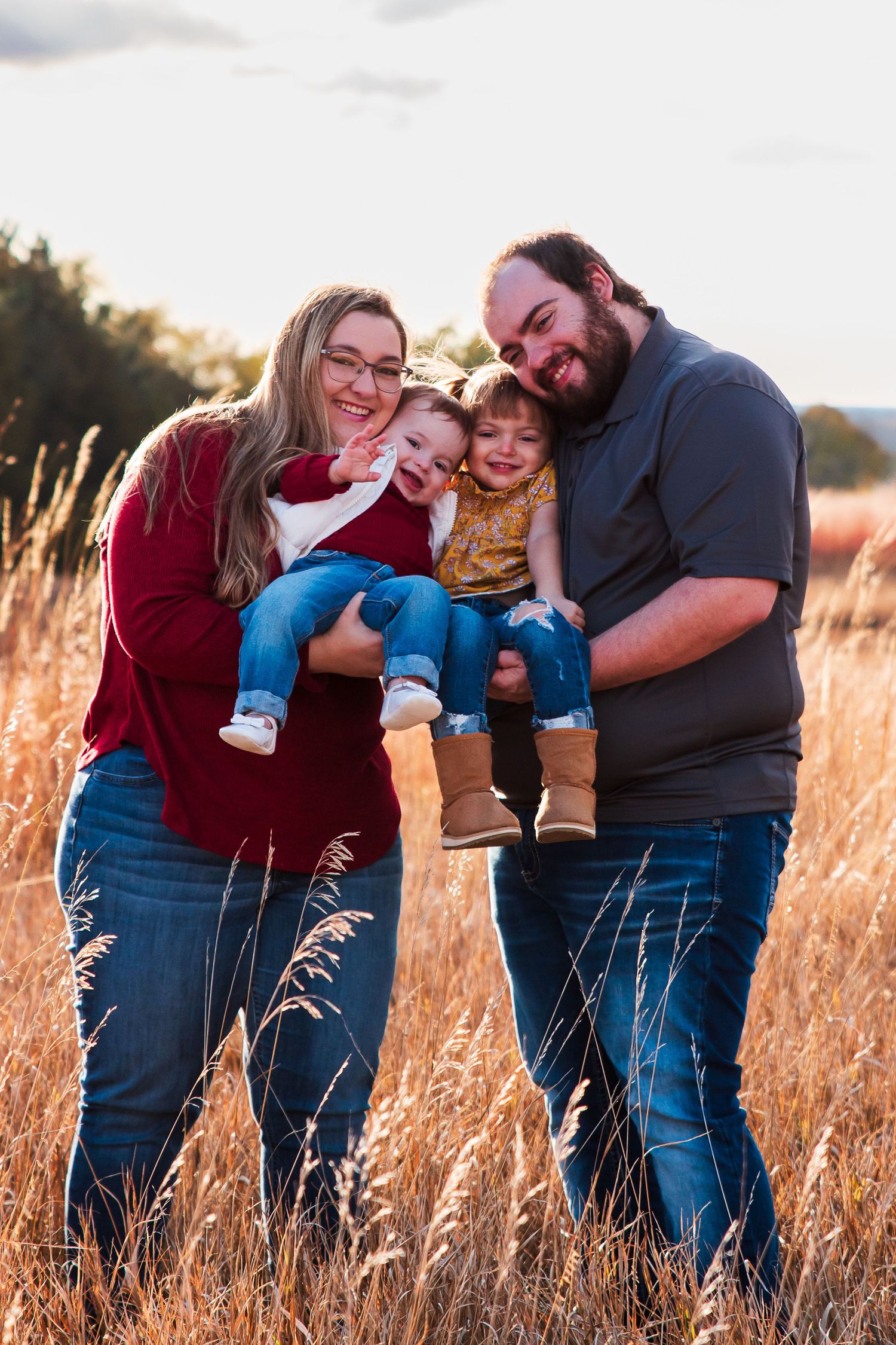 Family of four poses in golden field at sunset, parents holding two young children, smiling outdoors.
