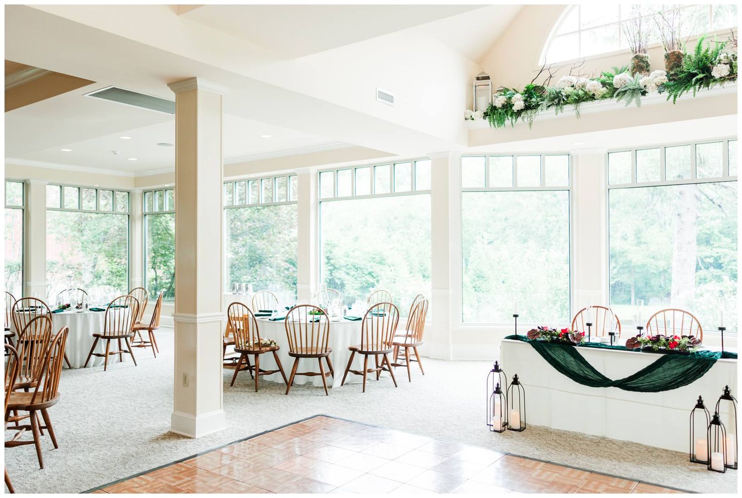 Bright wedding reception venue with large windows, round tables, wooden chairs, and green floral decor.