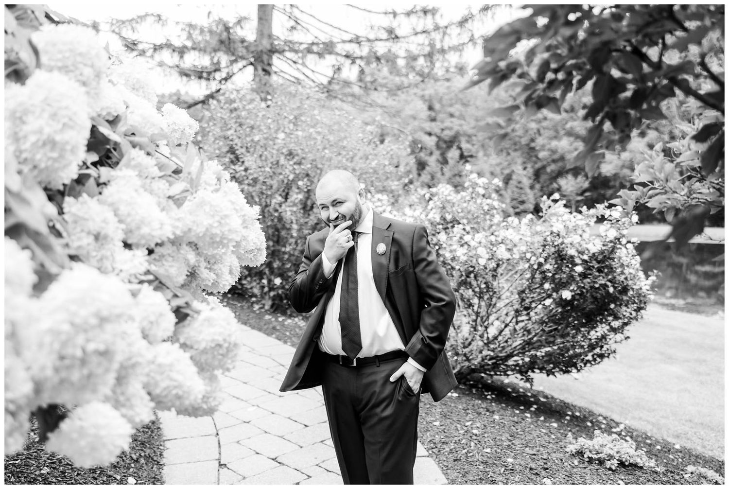 Elegant groom in black suit standing near white hydrangea bushes in black and white photo.