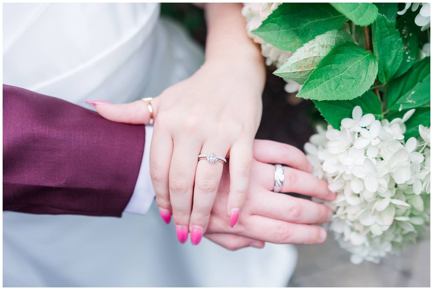 Close-up of bride and groom hands showing wedding rings with pink nails and white hydrangeas.