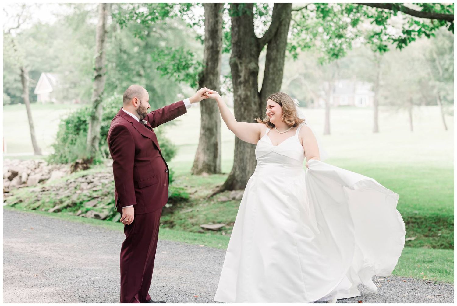 Bride and groom dancing outdoors, groom in burgundy suit twirling bride in white gown near lush green trees.