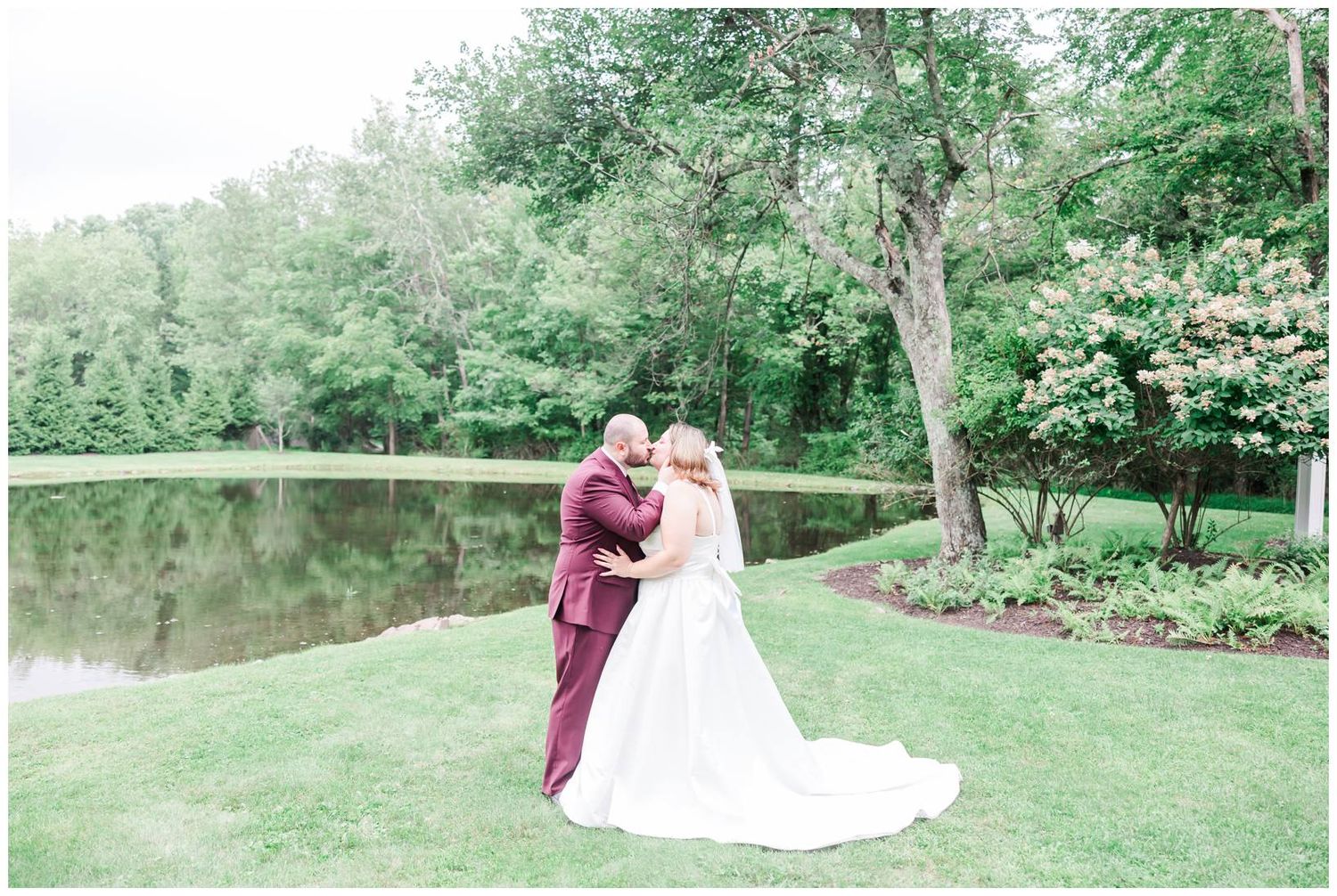 Bride and groom embrace romantically by a serene pond surrounded by lush greenery during outdoor wedding portraits.