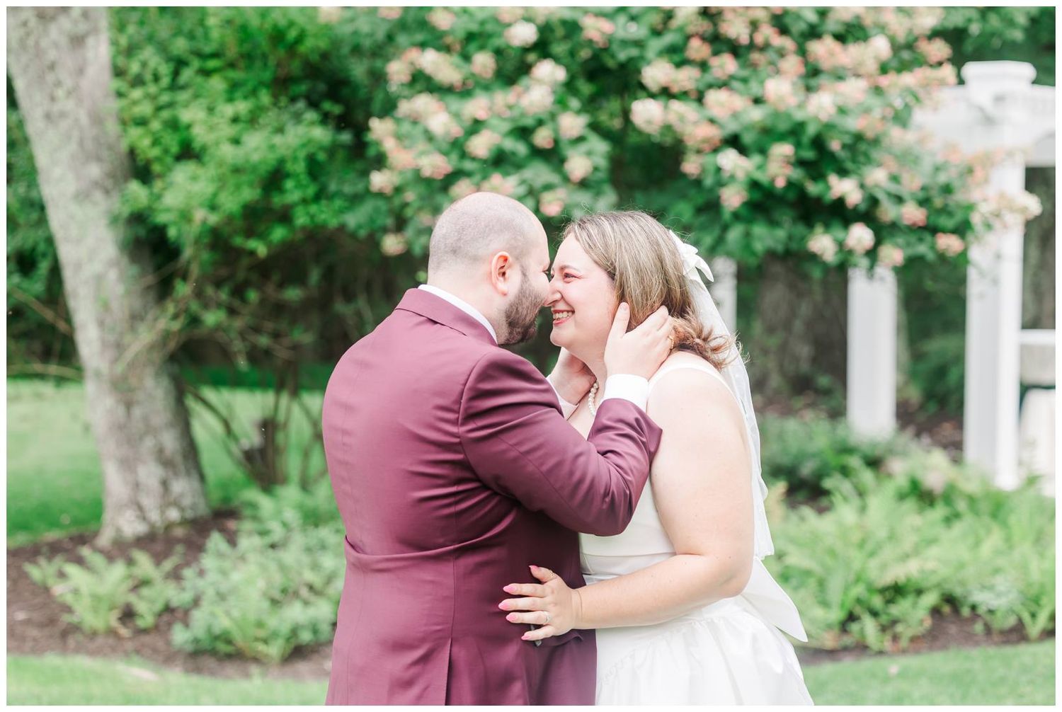 Bride and groom share a tender moment in lush garden, groom in burgundy suit gently cupping bride's face.