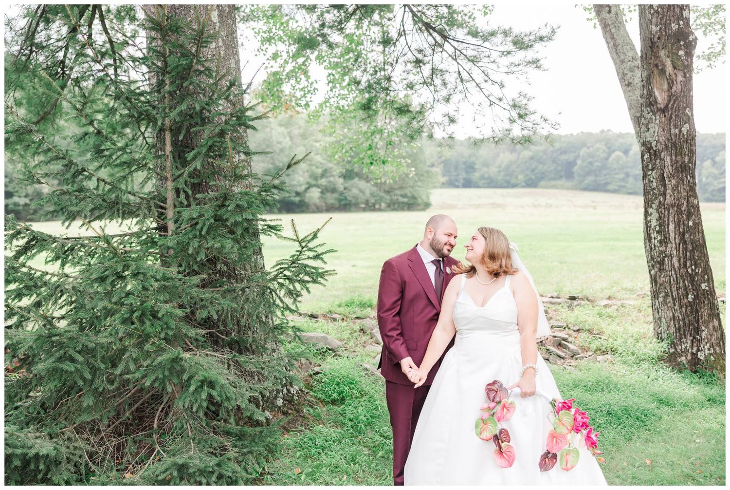 Bride and groom share a tender moment beside tall evergreen trees in a serene green countryside setting.