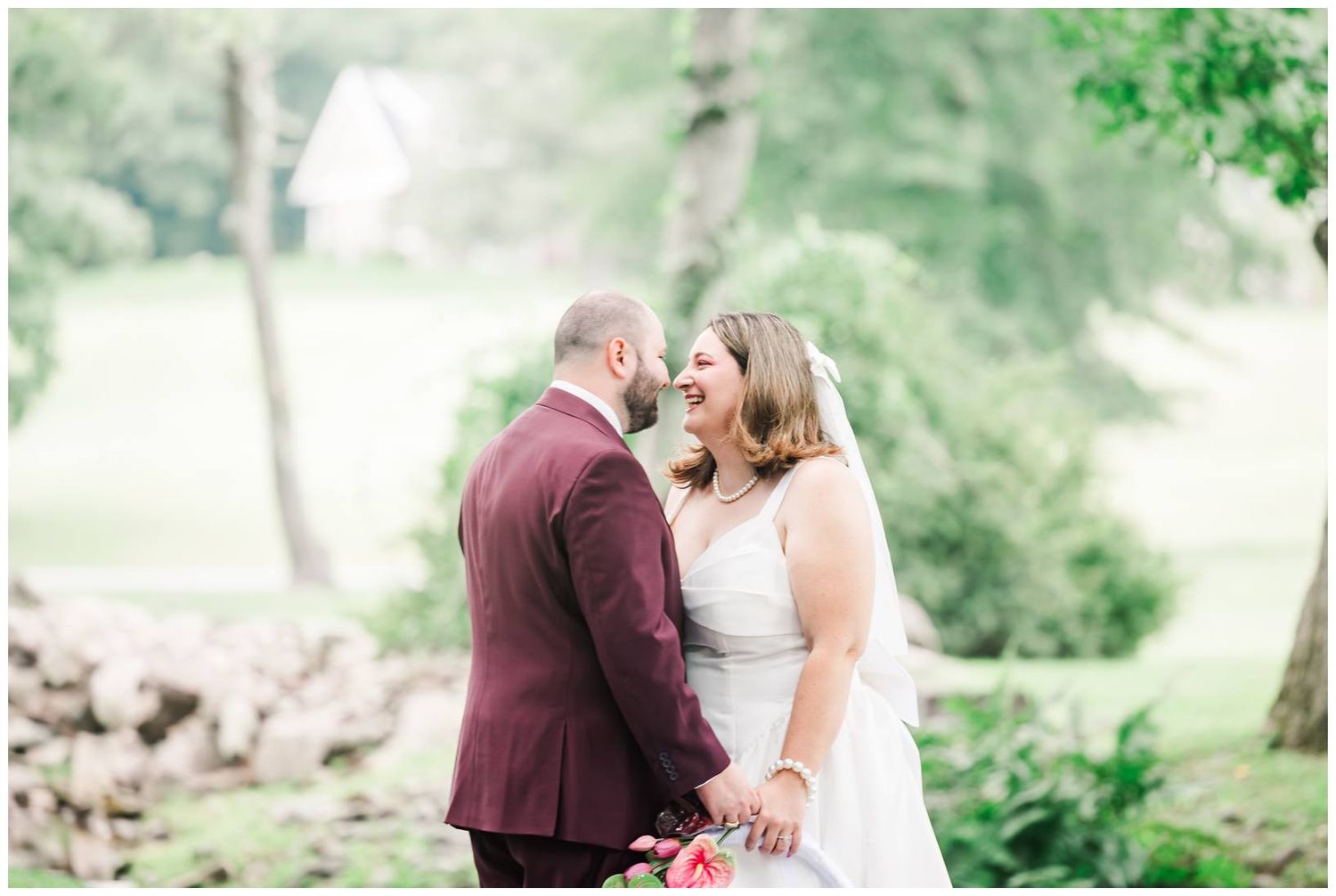 Bride and groom laughing together outdoors on their wedding day, surrounded by lush green trees.