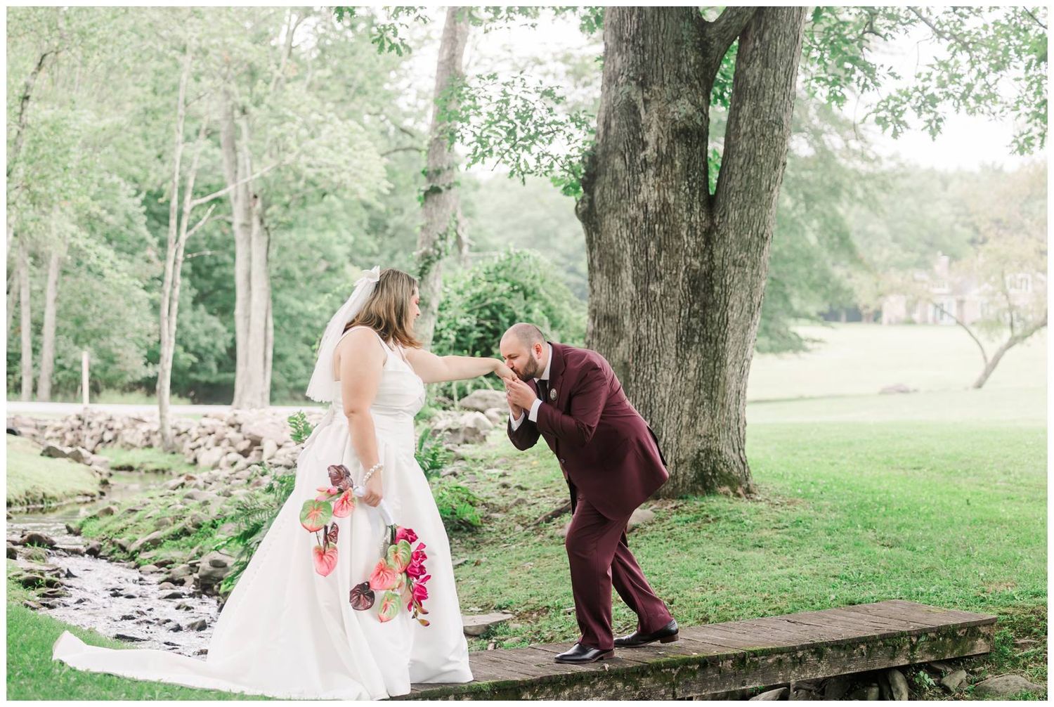 Groom in burgundy suit kisses bride's hand on rustic bridge surrounded by lush greenery at outdoor wedding.