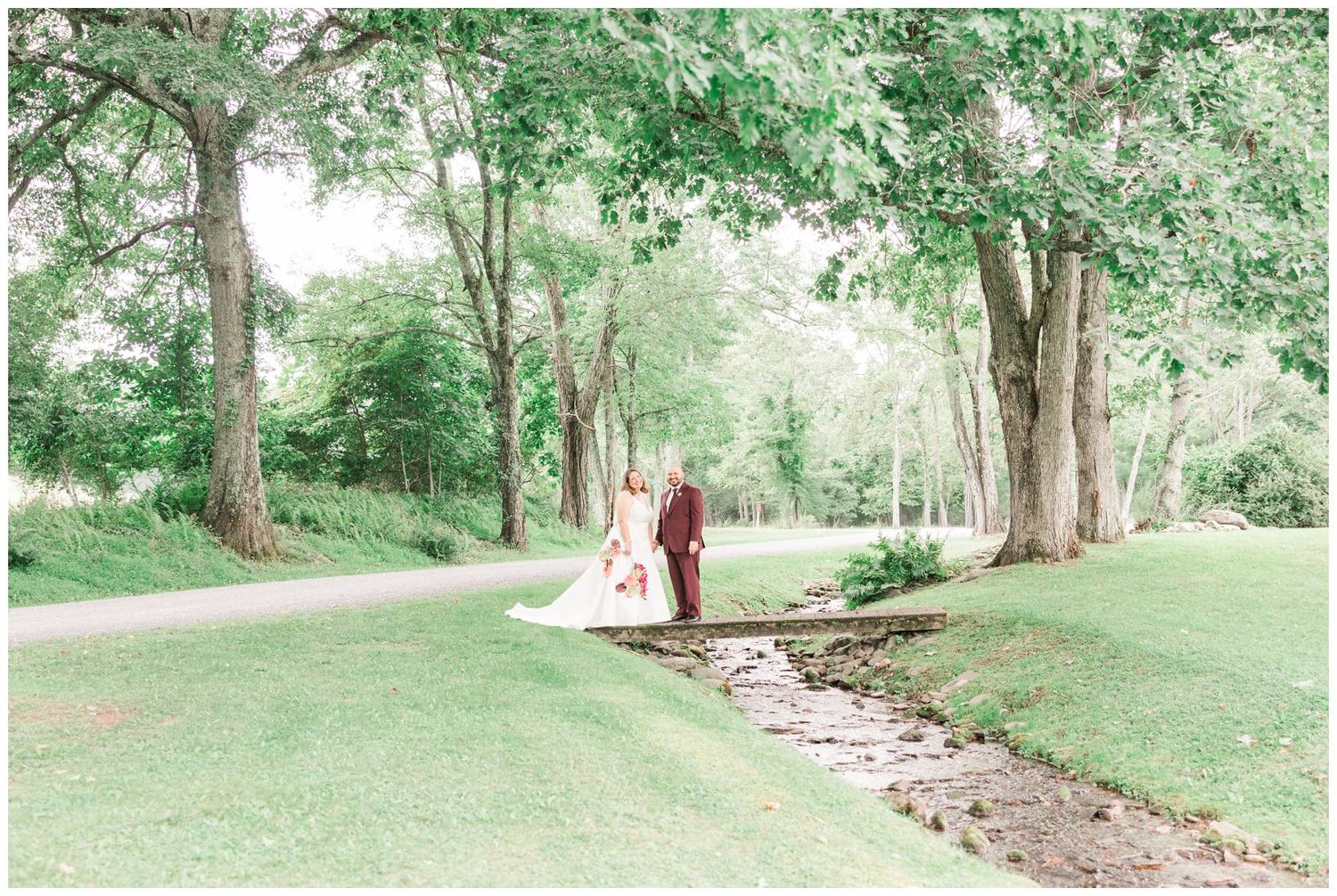 Bride and groom walk hand in hand across a lush green lawn beside a winding path surrounded by tall trees.