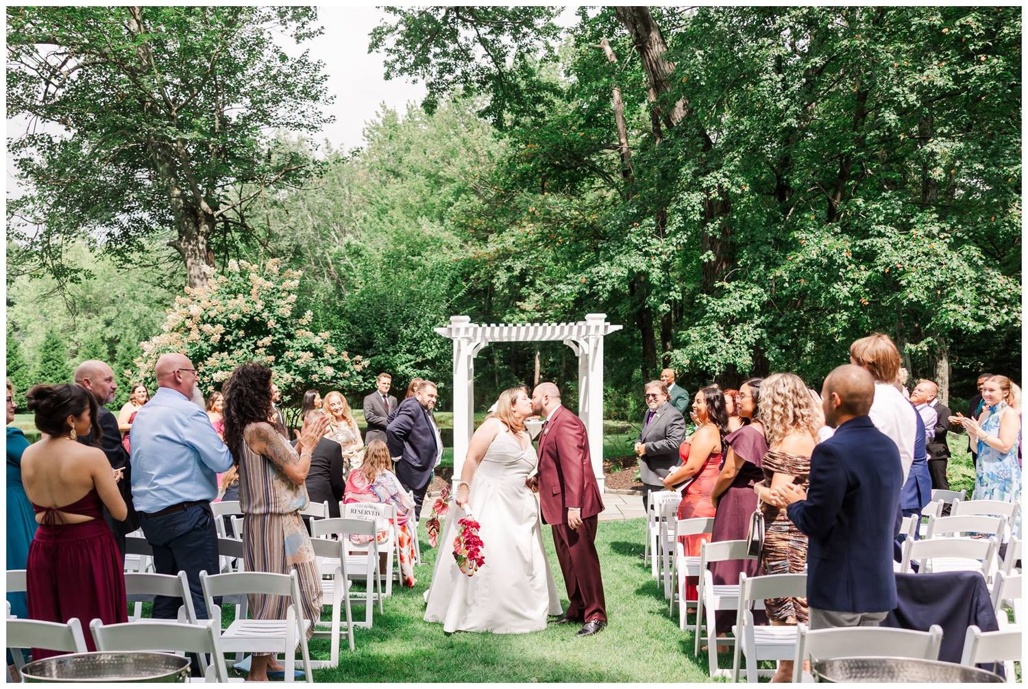 Bride and groom share first kiss as married couple while guests cheer at outdoor ceremony.