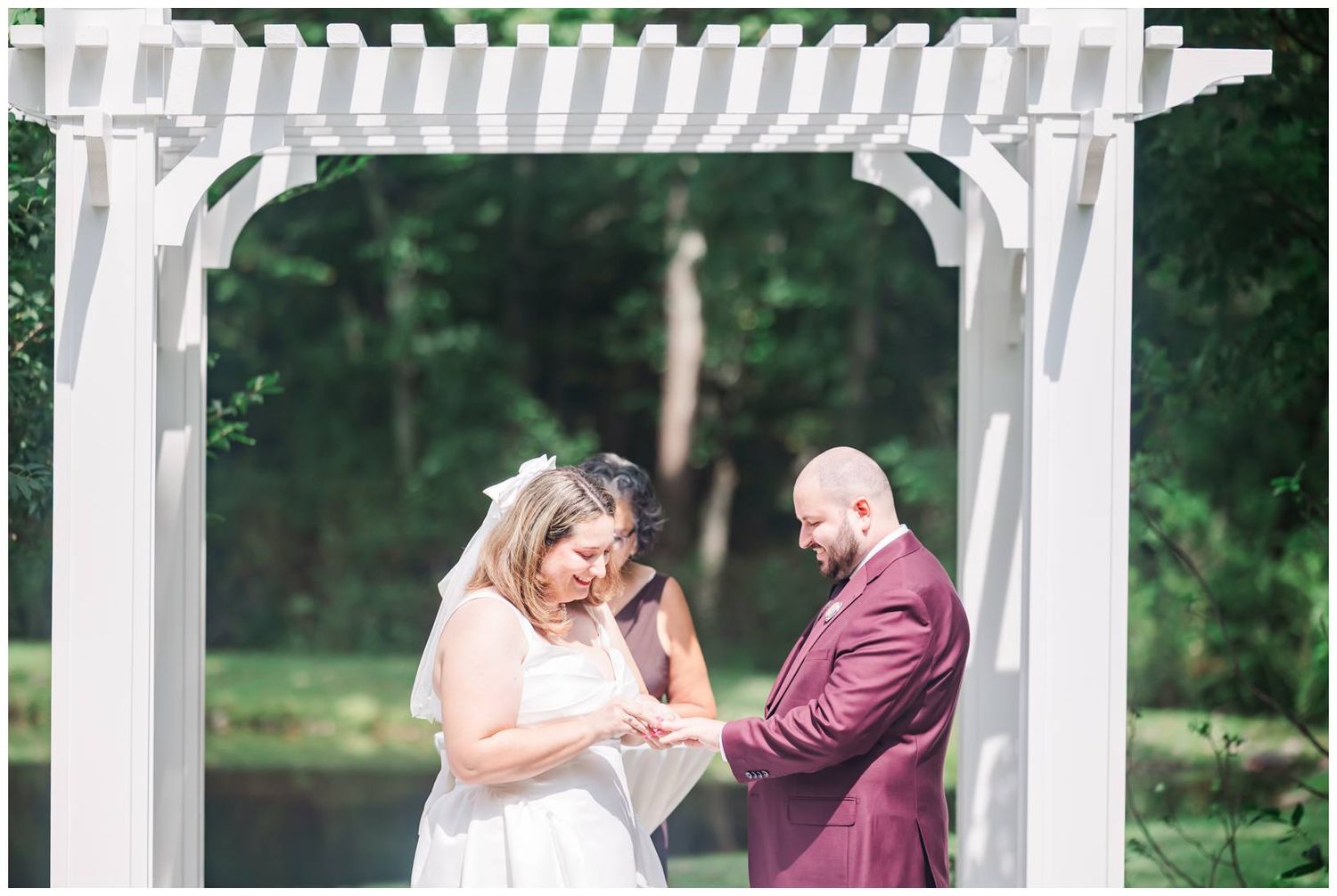 Bride and groom exchange rings under white garden arbor during outdoor wedding ceremony surrounded by lush green trees.