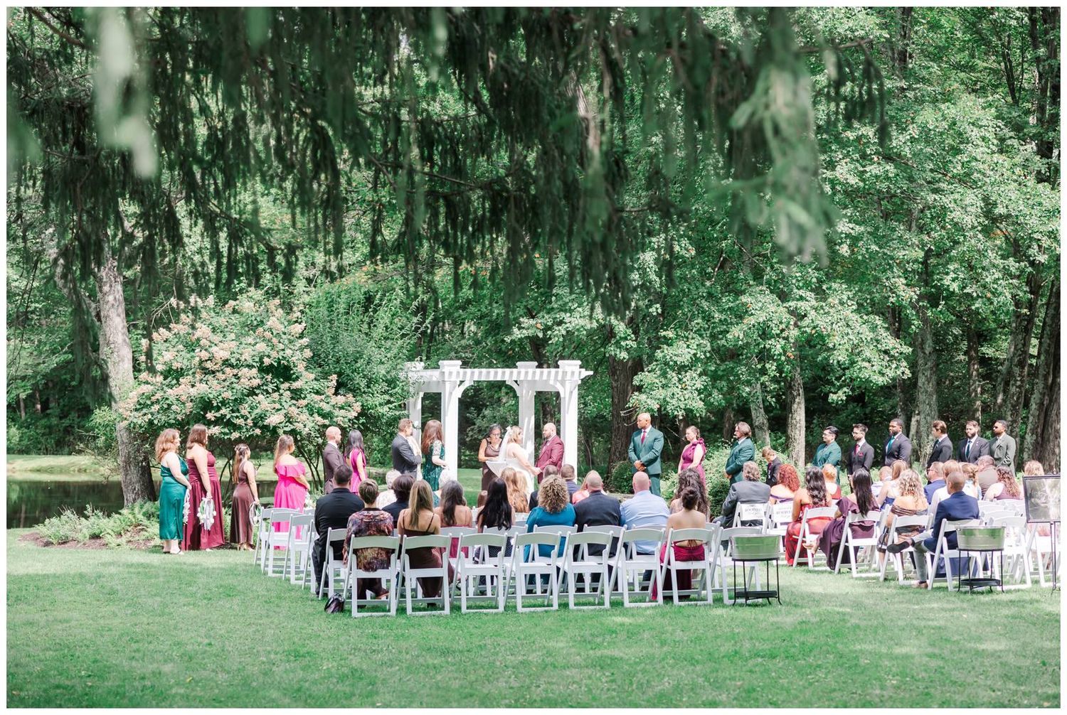 Outdoor wedding ceremony on green lawn with white arbor altar surrounded by lush trees and seated guests.