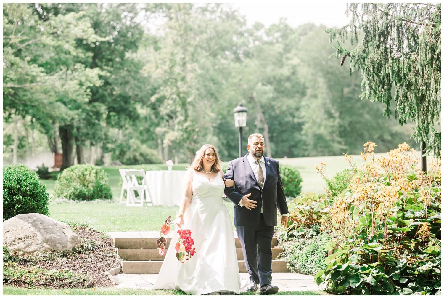 Bride in white gown with red bouquet walks with escort past lush garden greenery toward outdoor ceremony.