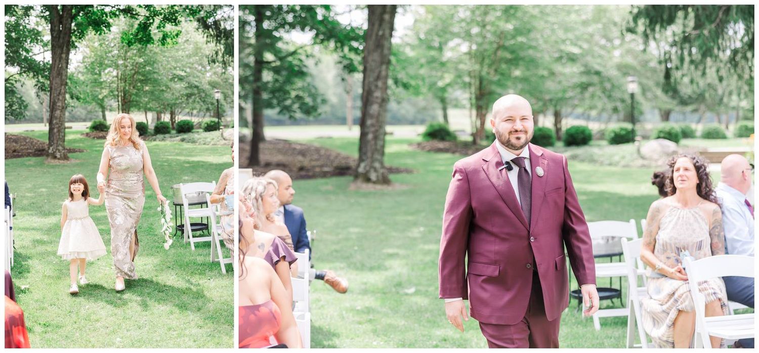 Groom in burgundy suit walks down outdoor wedding aisle flanked by guests seated in white chairs.