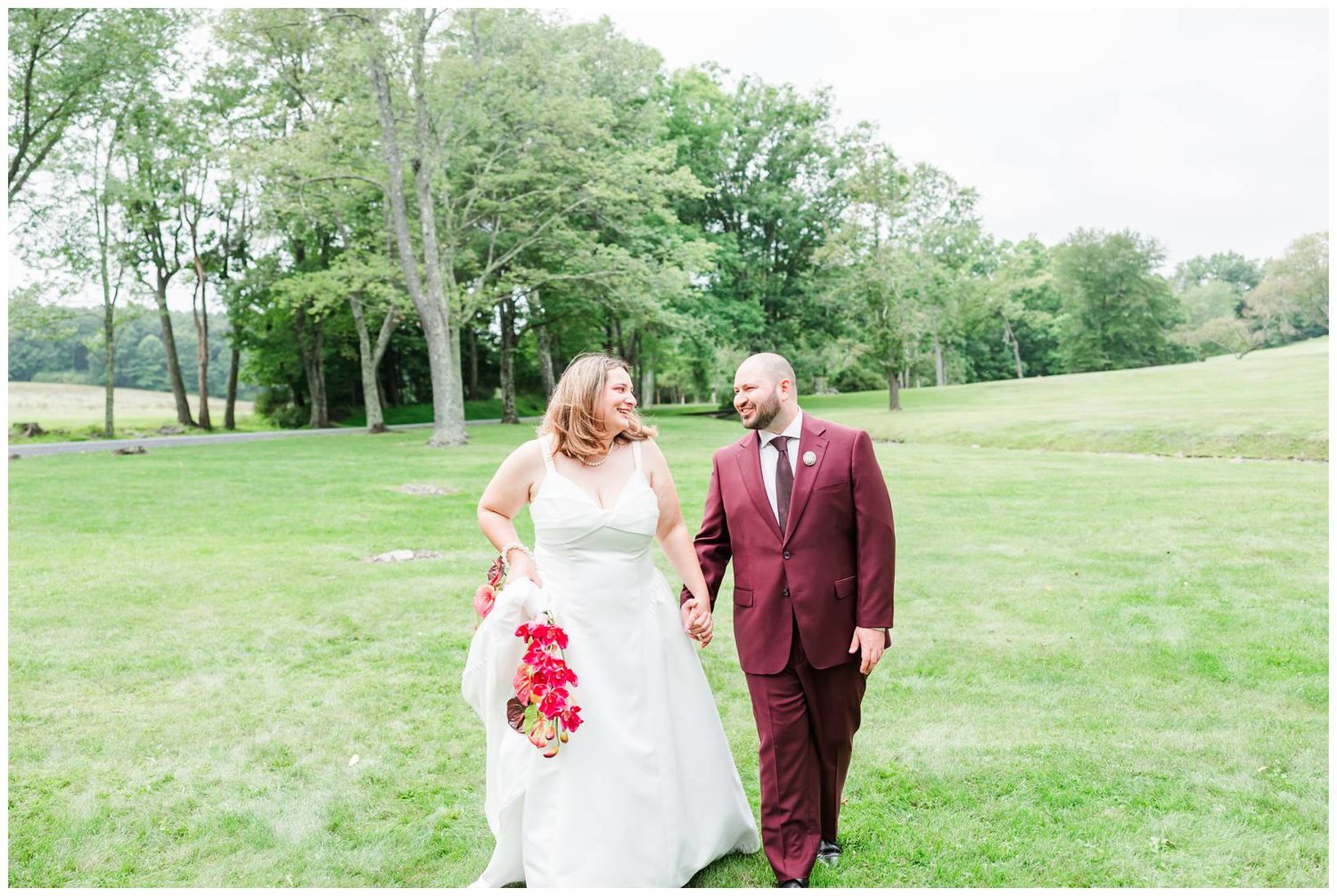 Smiling bride and groom walk hand-in-hand across green grass, groom in burgundy suit, bride in white gown.
