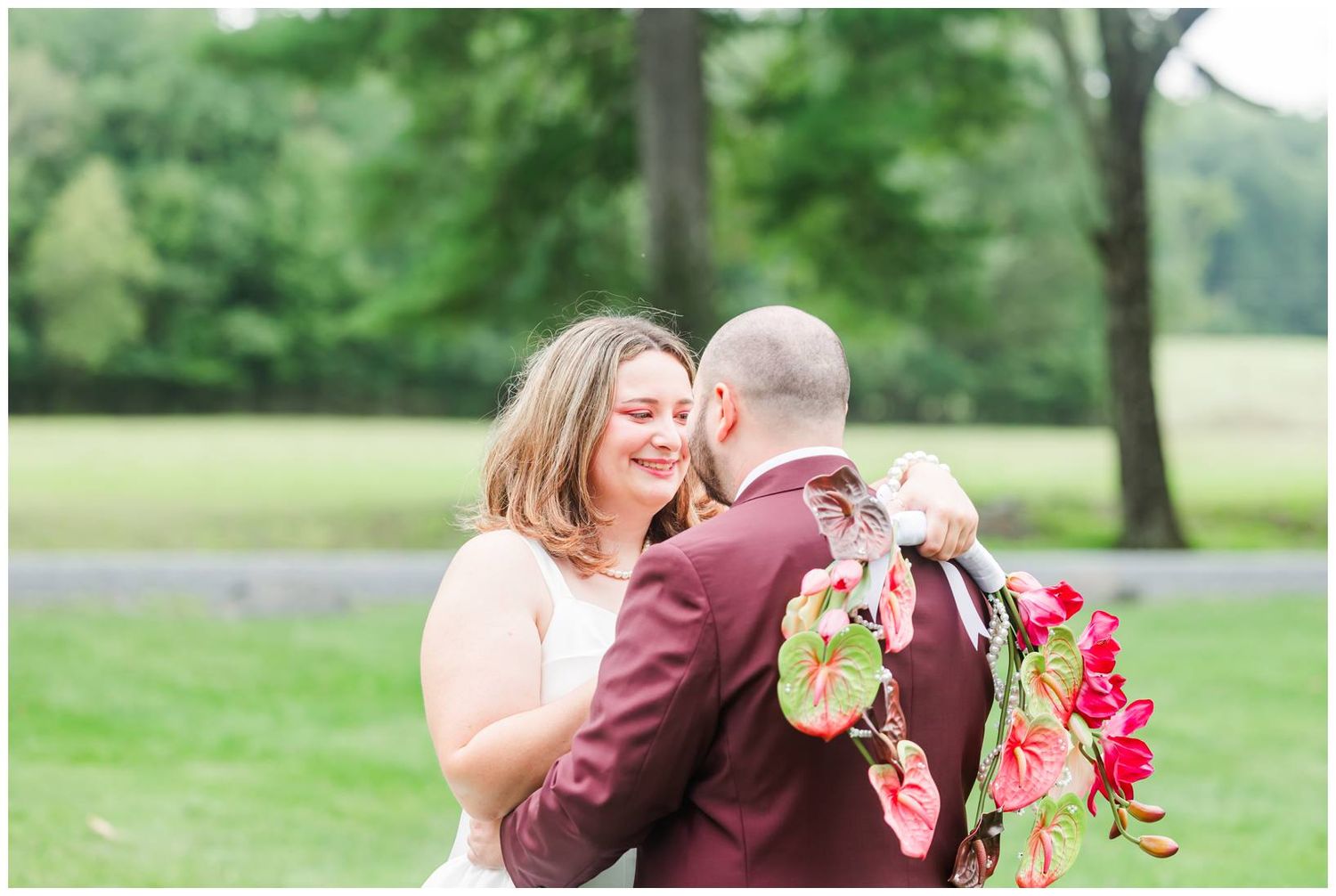 Bride and groom share tender embrace outdoors, groom in burgundy suit holding colorful bouquet.