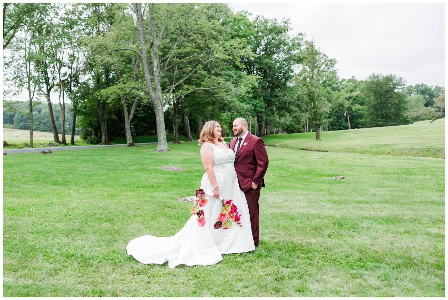 Bride and groom pose together on lush green lawn, groom in burgundy suit holding bride in white gown with red bouquet.