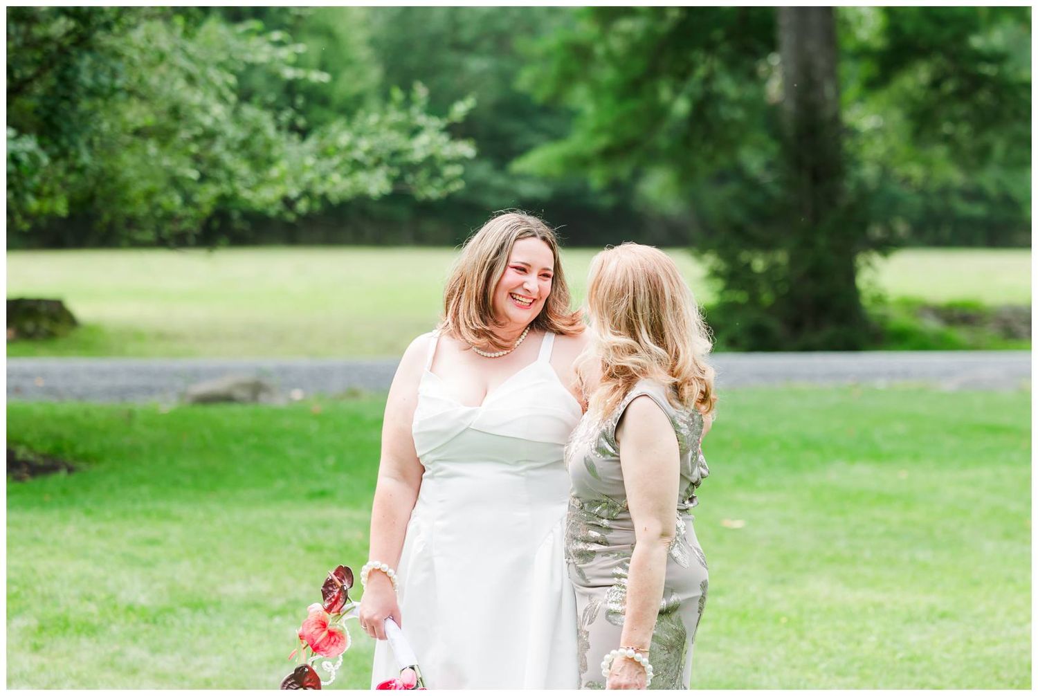 Two women in elegant attire share a joyful moment outdoors on a lush green lawn at a wedding celebration.