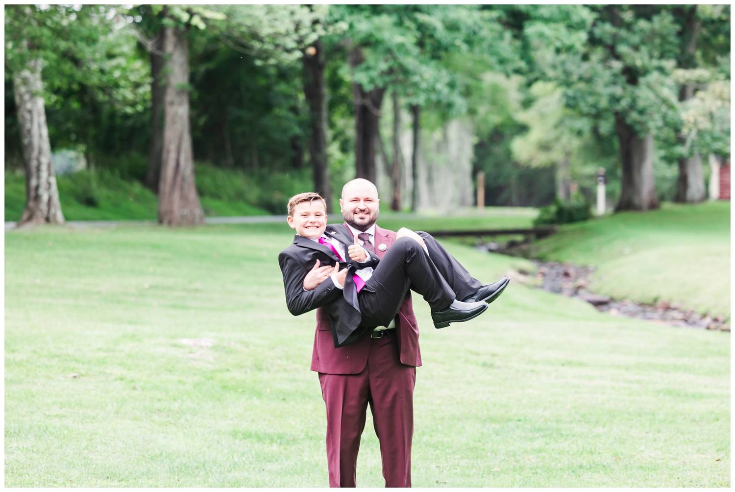 Groomsman in burgundy suit playfully carries another man in black suit at outdoor wedding.