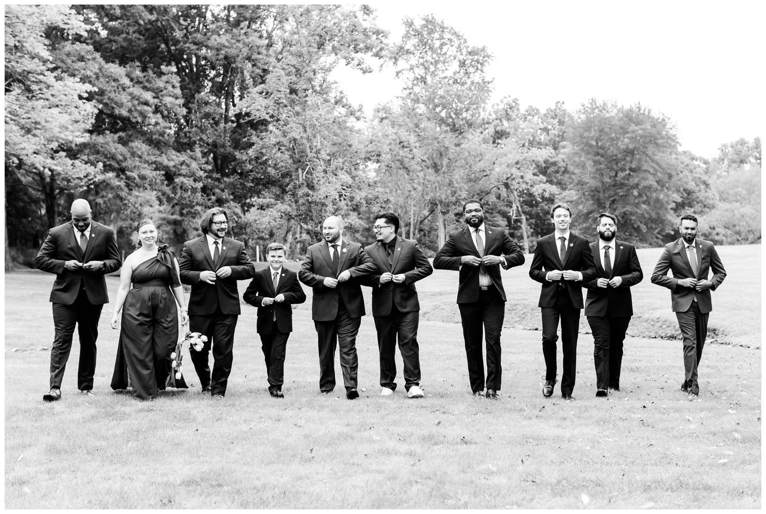 Wedding party in dark formal attire walking arm-in-arm across a grassy lawn with trees in the background.