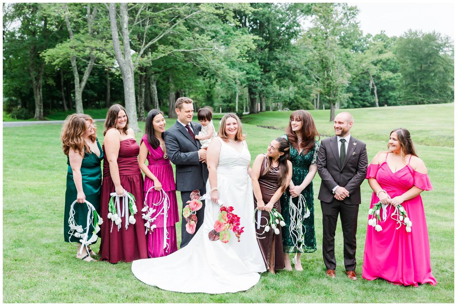 Wedding party poses on green lawn, bridesmaids in burgundy and pink gowns with groomsmen in dark suits.