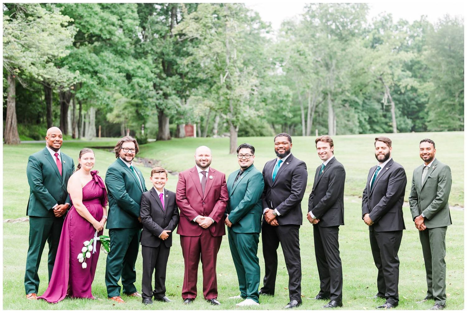 Groom in burgundy suit stands with groomsmen and bridesmaids in green and pink dresses on a lush lawn.