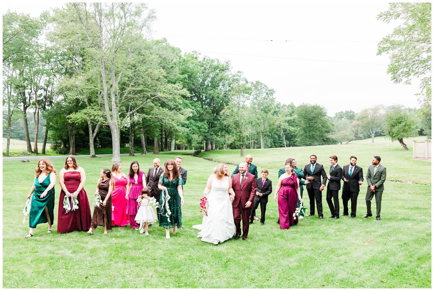 Wedding party in burgundy and green gowns with groom in burgundy suit on a lush green lawn.