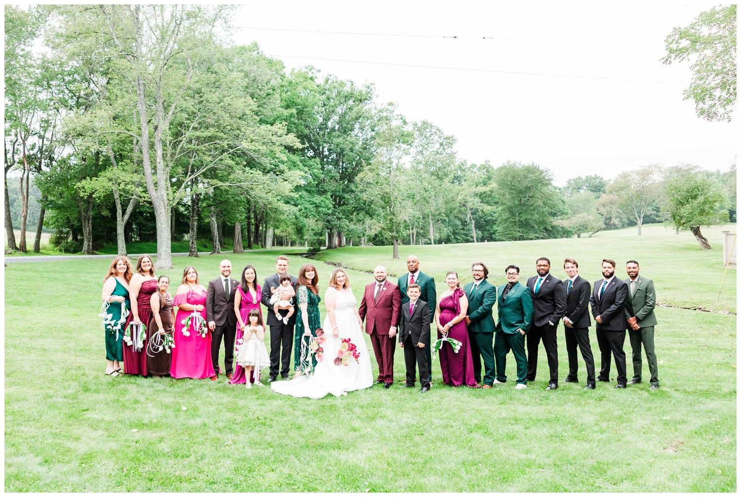 Wedding party in coordinating burgundy and green attire posing outdoors on lush green lawn surrounded by trees.