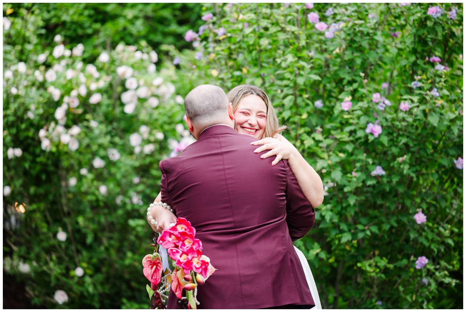 Couple embracing in a lush garden, groom in burgundy suit holding red bouquet, bride smiling joyfully.