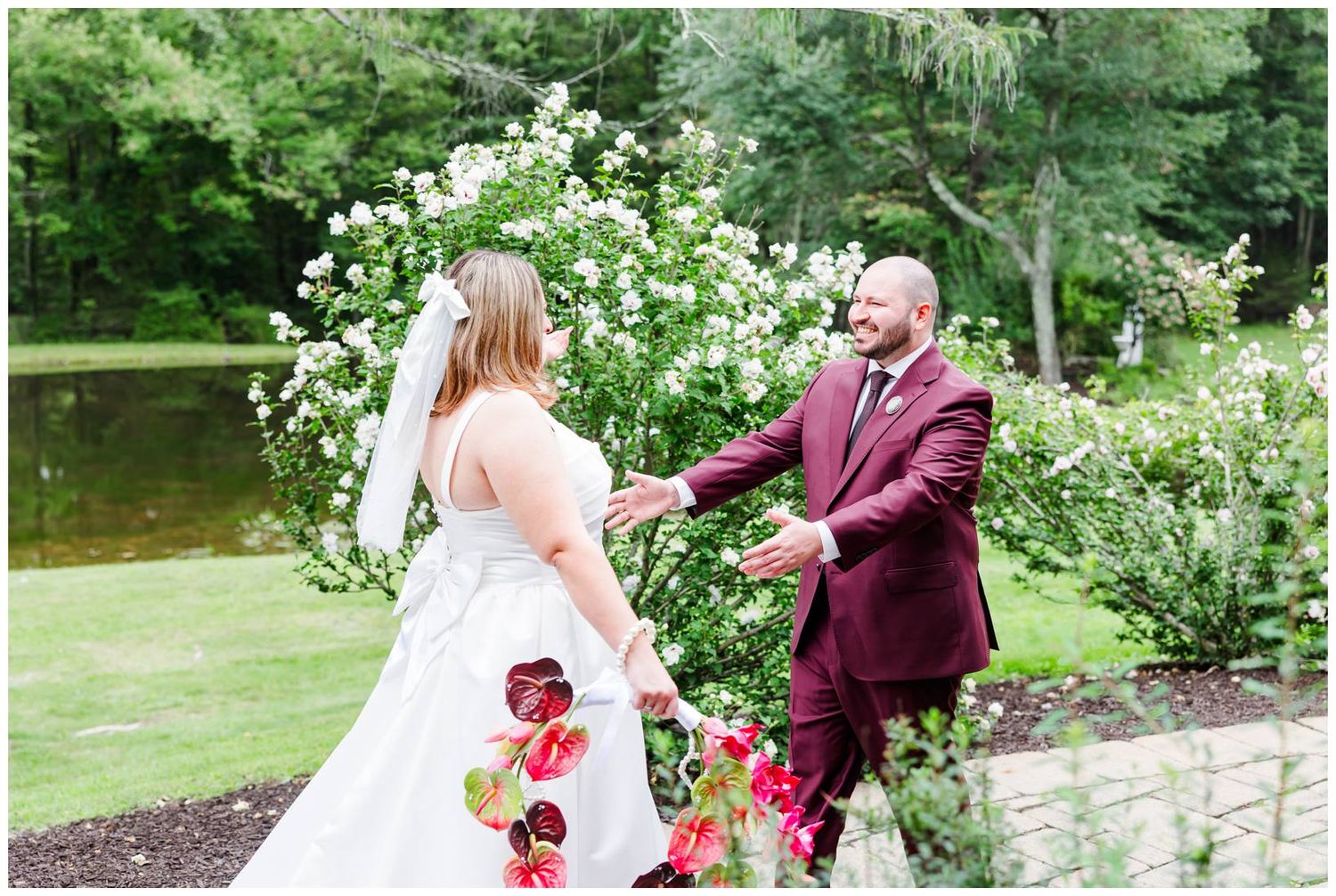 Bride and groom share a tender first look moment in a lush garden with white blooms and a pond backdrop.
