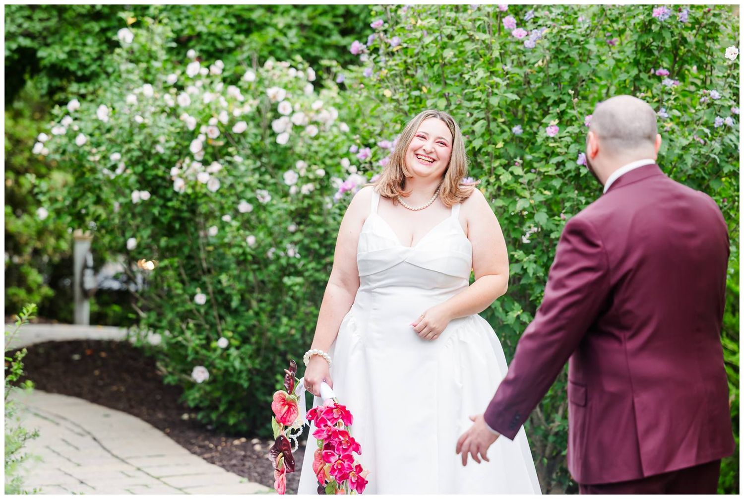 Bride in white gown smiles holding pink bouquet near blooming bushes during wedding first look.