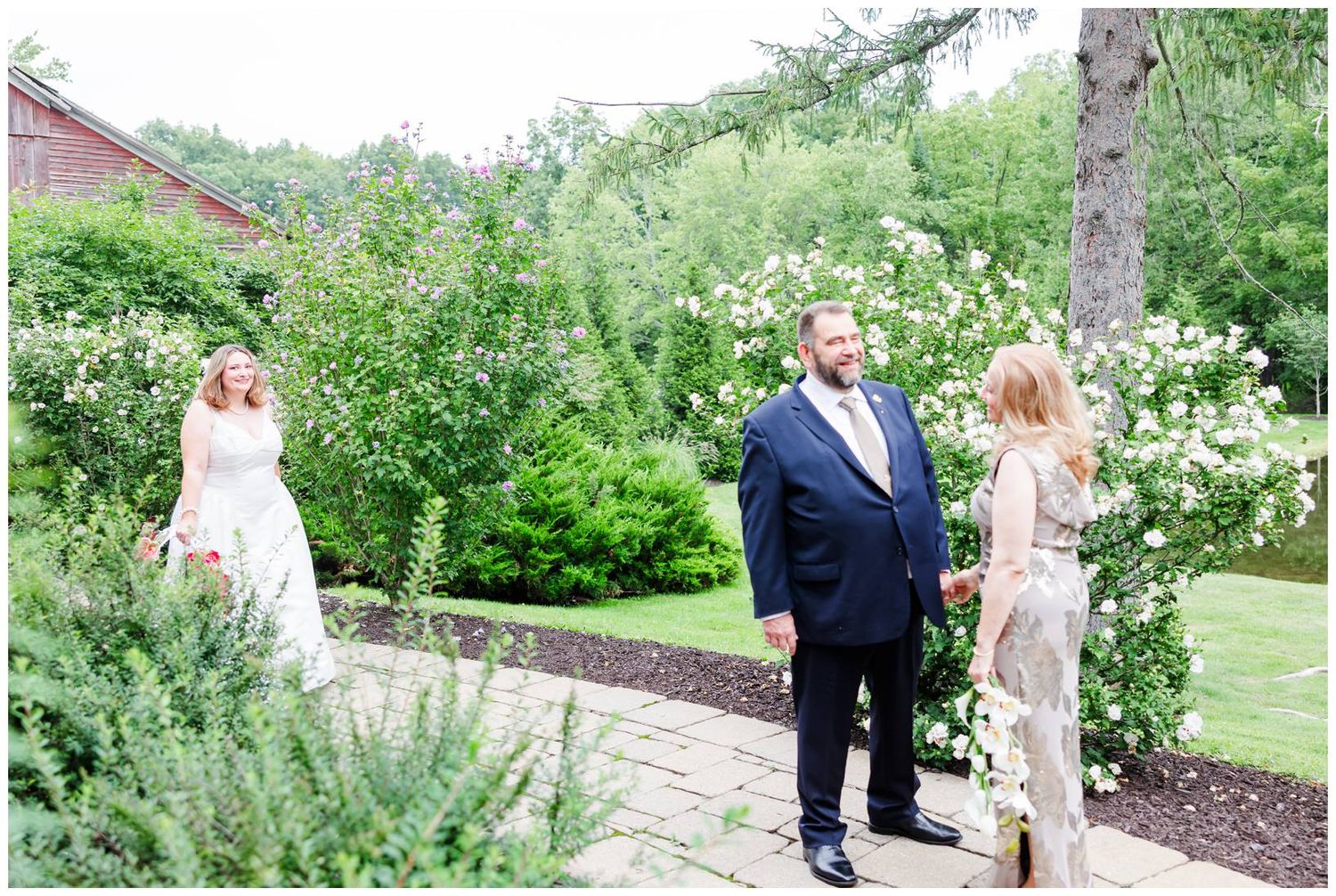 Groom in navy suit stands between two women in white dresses in a lush garden with blooming white flowers.