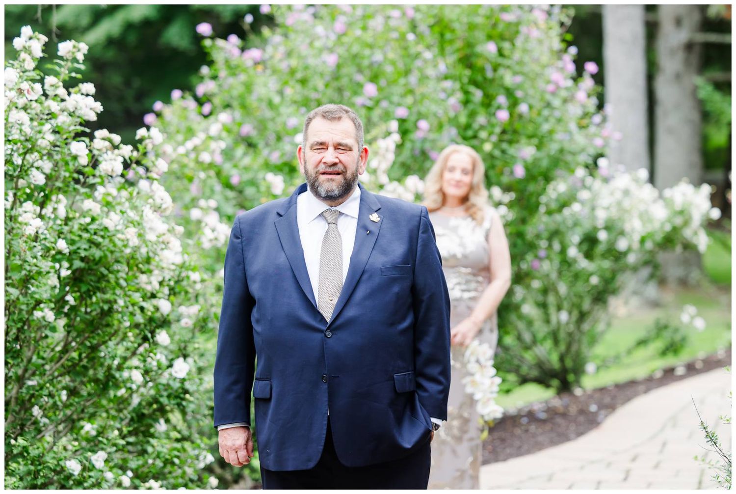 Groom in navy suit smiles during outdoor first look with bride among blooming white flowers.
