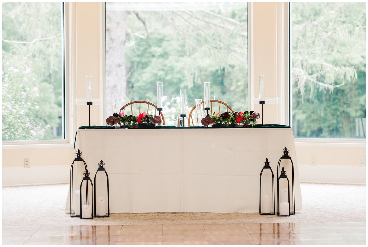Elegant wedding head table with white linen, red floral arrangements, and lanterns before large windows.