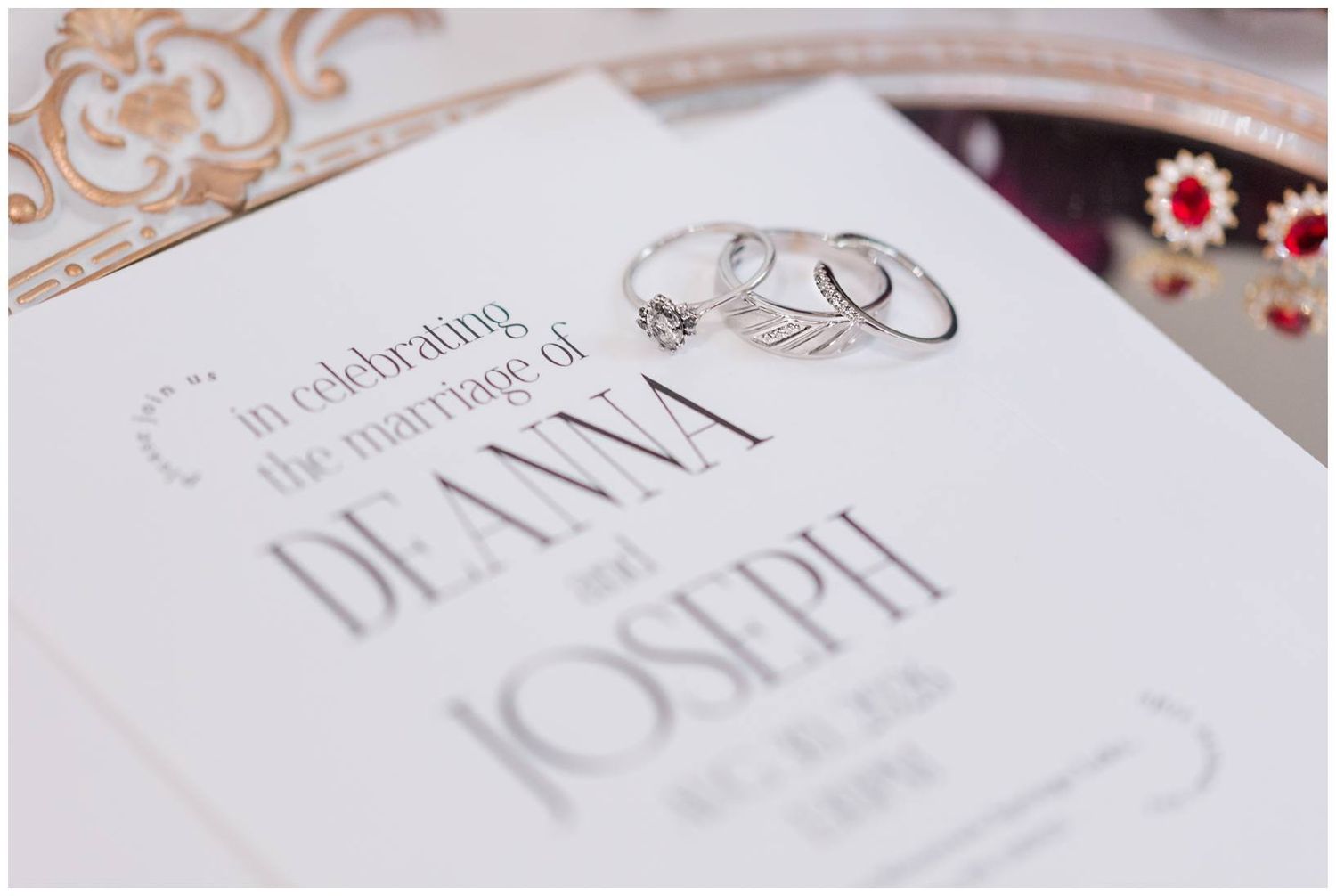 Wedding invitation for Deanna and Joseph displayed with elegant silver rings and ruby earrings on a white background.
