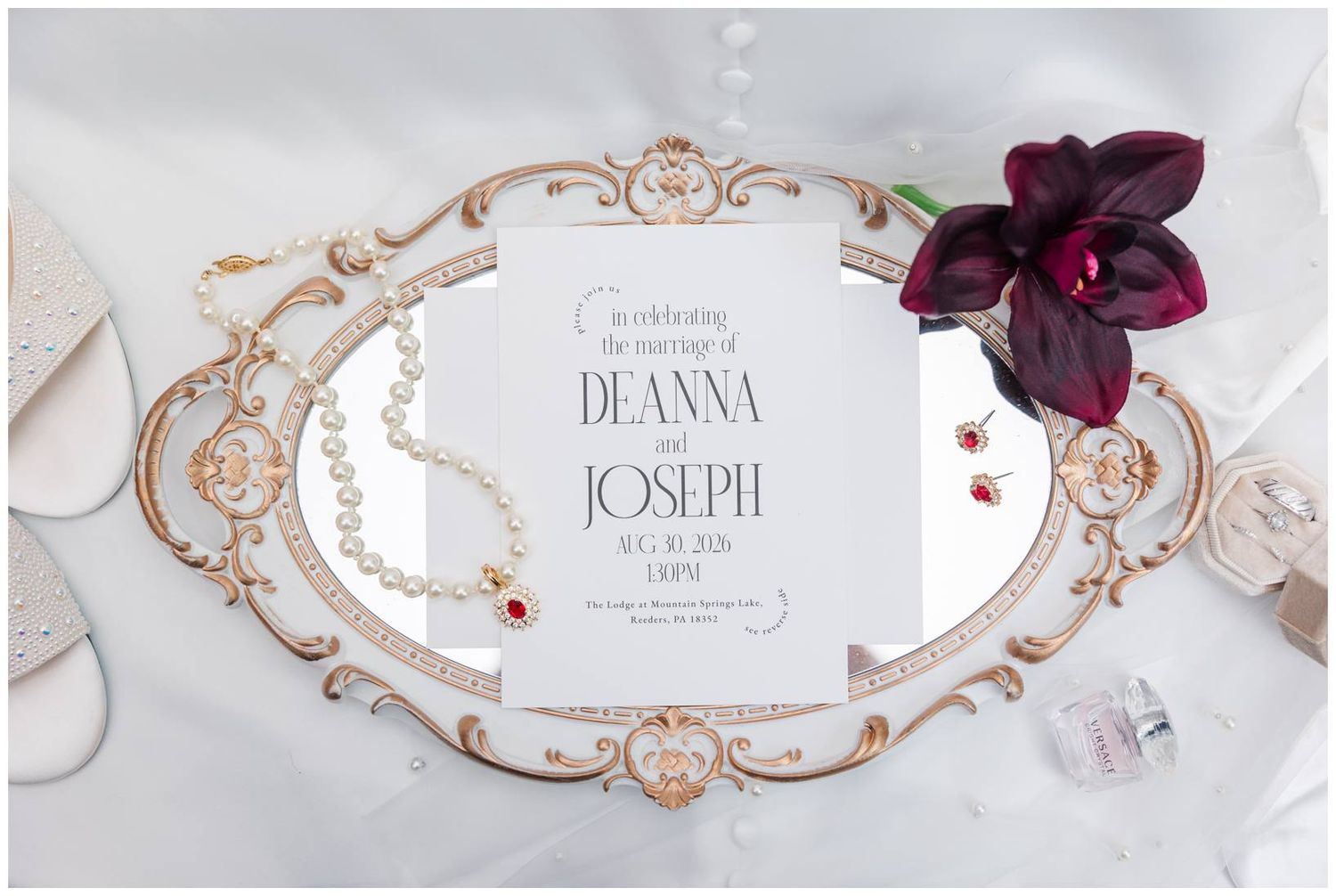 Wedding invitation for Deanna and Joseph displayed on ornate gold mirror with pearl bracelet, ruby earrings, and dark orchid.