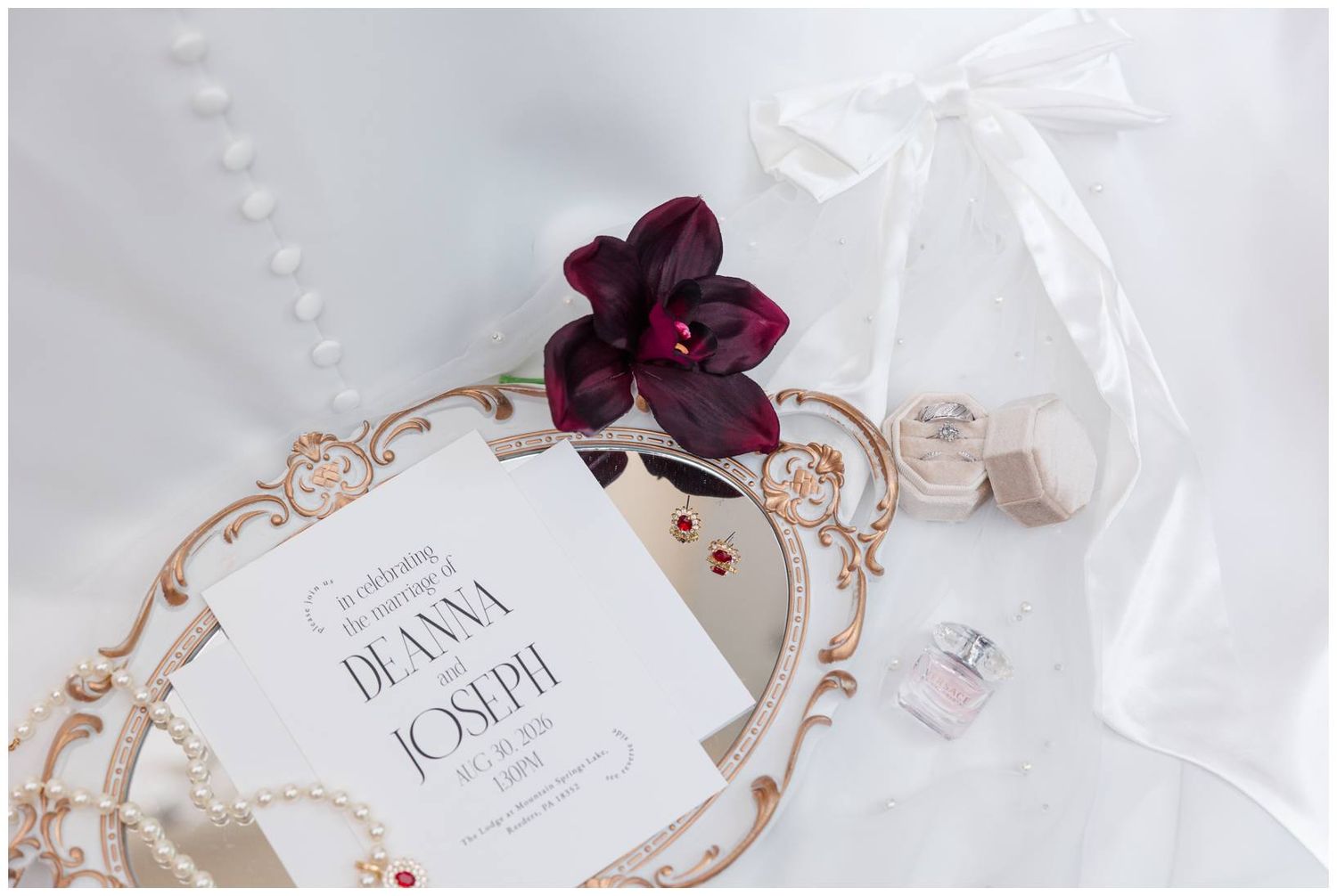 Elegant wedding invitation for Deanna and Joseph displayed on ornate tray with burgundy orchid, pearls, and perfume bottles.