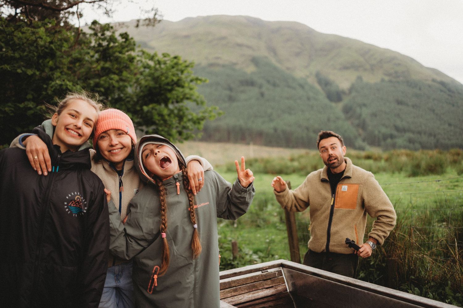 Four smiling people pose together outdoors with green mountains in the background on a cloudy day.